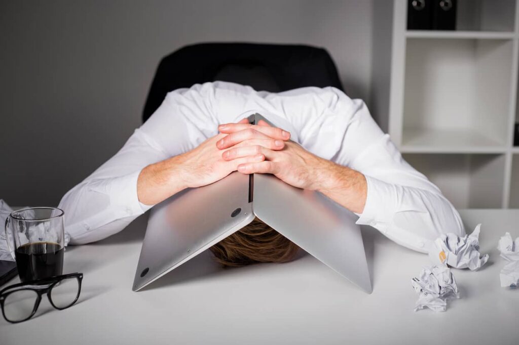 A man's face is pressed flat against the table, with a laptop covering the back of his head.